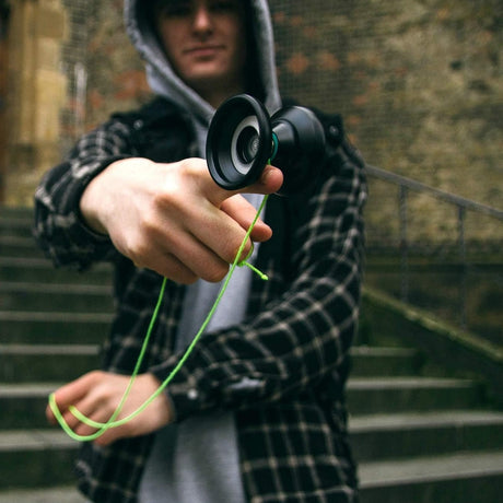 Närbild på en person i grå hoodie och rutig skjorta som balanserar en svart jojo på fingret med ett neon-grönt snöre. Personen står utomhus framför en stentrappa.