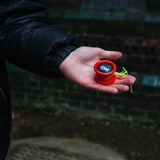 Närbild på en röd YoYoFactory One som ligger i en handflata utomhus. Jojon har en svart och blå logotyp med texten "ONE" och ett neongrönt snöre är lindat runt den.
