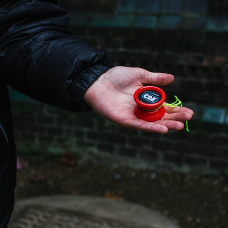 Närbild på en röd YoYoFactory One som ligger i en handflata utomhus. Jojon har en svart och blå logotyp med texten "ONE" och ett neongrönt snöre är lindat runt den.