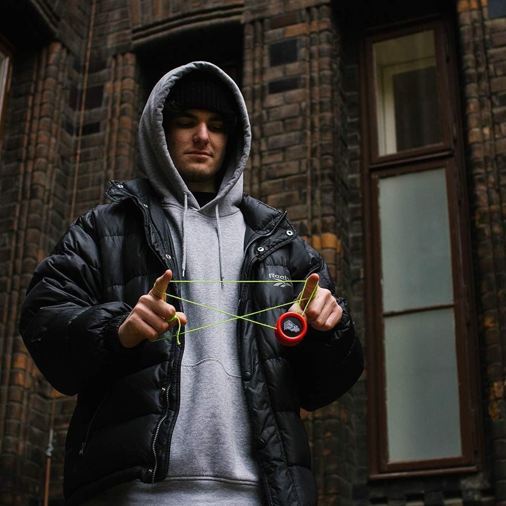 En ung man i grå hoddie och svart dunjacka utför ett avancerat yoyotrick med en röd Yoyo Factory One och ett neongrönt snöre utomhus mot en mörk tegelvägg